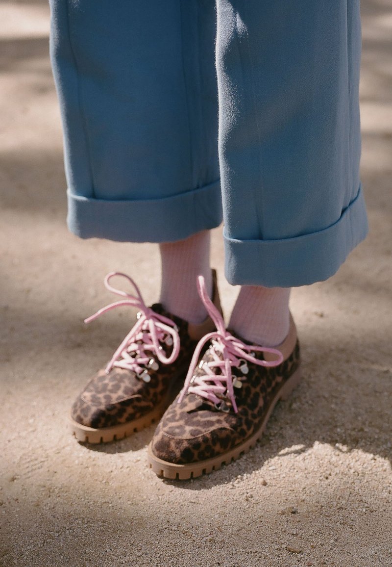 Personne portant un pantalon bleu et des chaussettes roses avec des chaussures marron à motif léopard et des lacets roses, debout sur une surface sableuse.