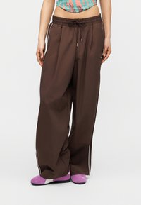 Άτομο που φοράει άνετο καφέ παντελόνι φόρμας με λευκές ρίγες στο πλάι και μοβ αθλητικά παπούτσια, στέκεται μπροστά σε ουδέτερο φόντο.