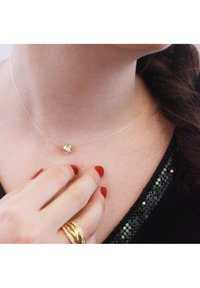Collana con pendente dorato e una pietra preziosa trasparente su una catena sottile e trasparente, indossata su un collo con smalto rosso visibile sulle dita.