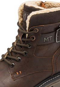 Botte en cuir marron avec doublure en fausse fourrure, bout arrondi, oeillets hexagonaux et couture orange contrastante. Détail de sangle réglable et de boucle.