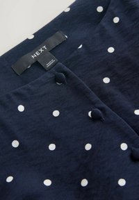 Mörkblå tygblus med vita prickar, som har rundad halsringning, knappar som detaljer och en etikett som visar märke och ursprung.