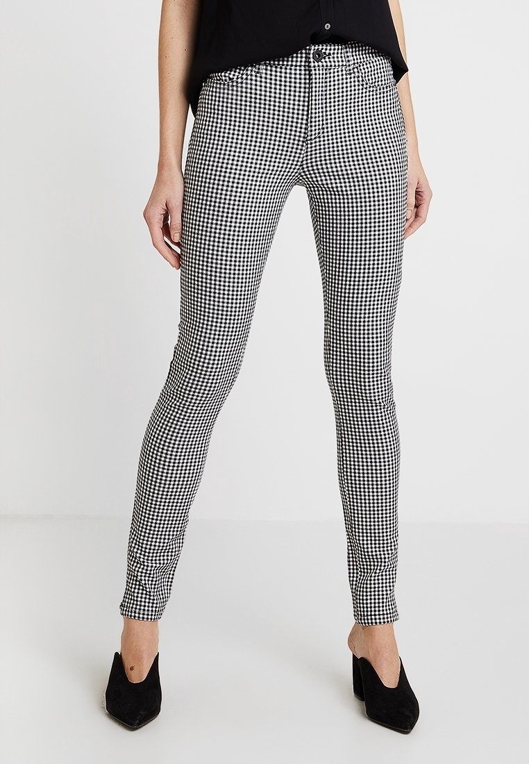 Femme portant un pantalon slim à carreaux noir et blanc, un haut noir sans manches et des chaussures à talons aiguilles noires pointues, debout sur un sol blanc.