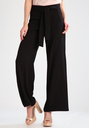 Vrouw draagt zwarte broek met hoge taille en wijde pijpen met een vooraan gestrikte riem en nudekleurige plateauzandalen met hoge hakken, staande tegen een effen achtergrond.