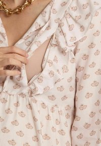 Kleid mit Blumenmuster aus leichtem pinkfarbenem Stoff, mit rüschigem Ausschnitt und einem Schlüssellochausschnitt. Es hat ein strukturiertes, mehrlagiges Design. Sichtbare goldene Kettenhalskette.