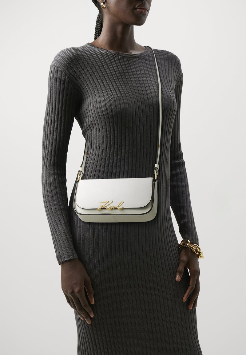 Sac en cuir blanc à bandoulière avec un logo doré, présentant un design épuré et des accents gris. Porté avec une robe grise à c�ôtes.