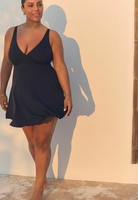 Schwarzes Badekleid mit tiefem V-Ausschnitt, schwingendem Rock und verstellbaren Trägern. Der glatte Stoff legt sich sanft über den Körper und betont die natürlichen Konturen.