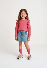 Rosa Langarmpullover mit Rüschenschultern, kombiniert mit einem hellen Denimrock mit used-Effekten und rosa knielangen Socken.