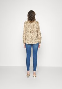 Blusa de estampado de serpiente beige con mangas largas, combinada con jeans ajustados azules y sandalias de tacón beige. El fondo es blanco liso.