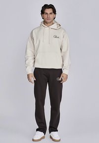 Beiger Kapuzenpullover mit Fronttasche und Kordelzug, mit besticktem Logo. Kombiniert mit dunkelbrauner Hose und weißen Sneakern. Einfaches Design.