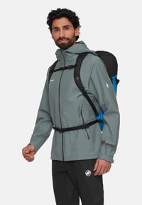 Ljust grått vattentätt jacka med huva, frontdragkedja, justerbara ärmslut och elastisk midja. Bärs över svarta byxor, med en blå ryggsäck.