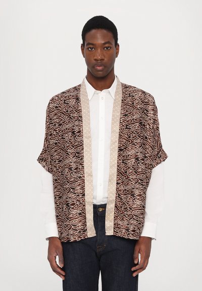 Brunmønstret kimono med hvit bølgedesign, med en kontrastfarge i beige langs kantene, båret over en hvit skjorte og mørke jeans.