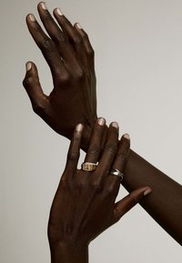 Dos manos con piel oscura, mostrando un anillo rectangular de oro y una banda de plata. Las uñas son naturales, de color claro, con textura y detalles visibles.