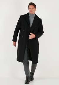 Cappotto nero in lana con design doppiopetto, grandi bottoni e revers a lancia, indossato sopra un dolcevita grigio e pantaloni grigi.