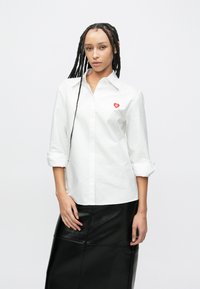Bijela košulja s gumbima i podvrnutim rukavima te crvenim logotipom srca na prsima, u kombinaciji s crnom kožnom suknjom. Glatka tekstura tkanine.