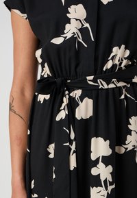 Melna kleita ar krēmveida ziedu rakstu, elastīgu jostu un sasienamu jostu. Gluda tekstūra, īsas piedurknes un plūstošs dizains.