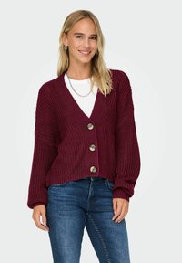Burgundská pletená vesta s výstřihem do V, velkými knoflíky a dlouhými rukávy. V kombinaci s bílým topem a modrými džíny, které mají volný střih.