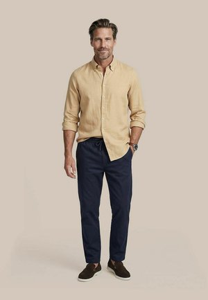Man staat, draagt een beige overhemd met knopen en opgerolde mouwen, marineblauwe broek met trekkoord, bruine instapschoenen en een polshorloge.
