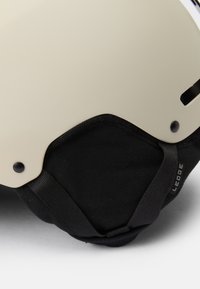Sigurnosna kaciga s mat krem bojom, crnim jastučićima i teksturiranim bočnim remenom s detaljem brendiranja.