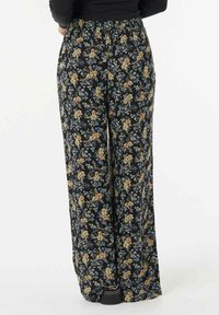 Personne portant un pantalon large à fleurs noirs et des chaussures noires, se tenant contre un fond blanc.