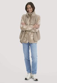 Beige fleece-takki, jossa on kukkakuosi, korkea kaulus ja vetoketjukiinnitys. Yhdistetty vaaleansinisiin farkkuihin ja neutraaleihin lenkkareihin. Rentoa tyyliä.