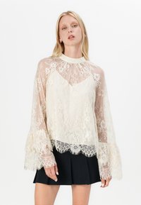 Camicia trasparente avorio con sovrapposizione in pizzo floreale, colletto alto e maniche a campana. La gonna nera plissettata completa l'outfit, con un intricato bordo in pizzo.