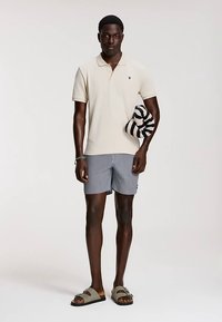 Bézs színű galléros póló, csíkos rövidnadrág, szürke szandál, fekete-fehér csíkos kiegészítőt tart. A póló mellrészén logó található.