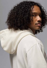 Huppari pehmeästä kermanvärisestä kankaasta, rento istuvuus, näkyvät ompelusaumat ja kiinnitetty huppu.