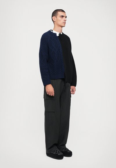 Knitted cardigan with a navy and black textured design. Features a mix of patterns. Paired with loose black cargo pants and patterned shoes.