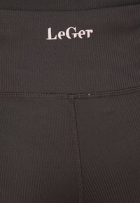 Μαύρα ριμπ κολάν με ψηλή μέση που φέρει το όνομα της μάρκας "LeGer" σε ανοιχτό ροζ. Ορατές ραφές κατά μήκος των πλευρών.