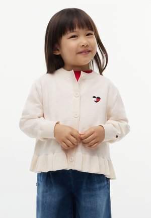 Jong kind glimlacht, draagt een crèmekleurige cardigan met knopen, gerimpelde zoom en geborduurd hart, en een blauwe spijkerbroek tegen een witte achtergrond.