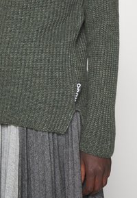 Ciemnoskóra ręka w zielonym swetrze z dzianiny z metką Hugo, w zestawieniu z szarym plisowanym spodnicą w wzór jodełki.