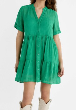 Femme portant une robe verte à manches courtes boutonnée, avec une jupe à volants et des manches retroussées, associée à des bottes de couleur claire.