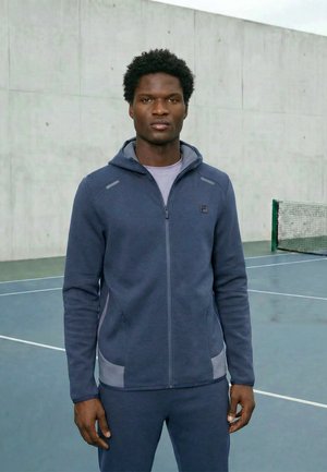 Noor mees, kes kannab tumelasinist lukuga kapuutsi ja dressipükse, seisab välises tennisekohas betoonseinaga taustal.