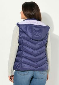 Granatowa kamizelka puchowa z kapturem, z wzorem w zygzak i jasnopurpurową podszewką, noszona na szarej koszulce z długim rękawem.