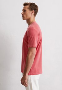 Short-sleeve coral t-shirt made of cotton, featuring a crew neck, relaxed fit, and subtle side tag detail, paired with white trousers.