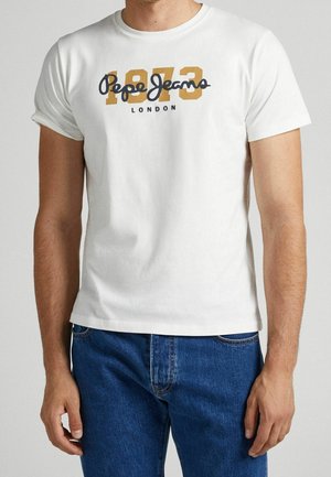 Mann trägt ein weißes Kurzarm-T-Shirt mit der Aufschrift „1983 Pepe Jeans London“ in Gelb und Schwarz, kombiniert mit blauen Jeans.