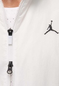 Chaqueta blanca con cremallera, con un logo Jumpman negro en el pecho, herrajes de la cremallera negros y un diseño de tela texturizada.