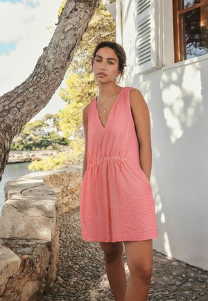 Junge Frau mit dunklen Haaren in einem ärmellosen korallenrosa Kleid, die an einer Steinmauer neben einem Baum und einem weißen Gebäude in einer sonnigen Außenanlage steht.
