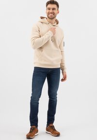 Felpa beige con tasca frontale e cappuccio con cordino, abbinata a jeans in denim scuro e scarpe da ginnastica marroni con lacci.