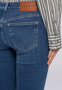 Acercamiento de unos pantalones vaqueros de denim azul con el bolsillo trasero y una etiqueta de la marca Trussardi en marrón, combinados con el puño de una camisa de rayas de manga larga y la mano descansando sobre el muslo.