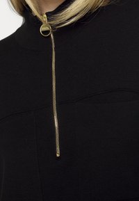 Close-up van een zwart gebreide top met een gouden rits en een ronde gouden rits-ritssluiting bij de hals, gedragen door een persoon met blond haar.