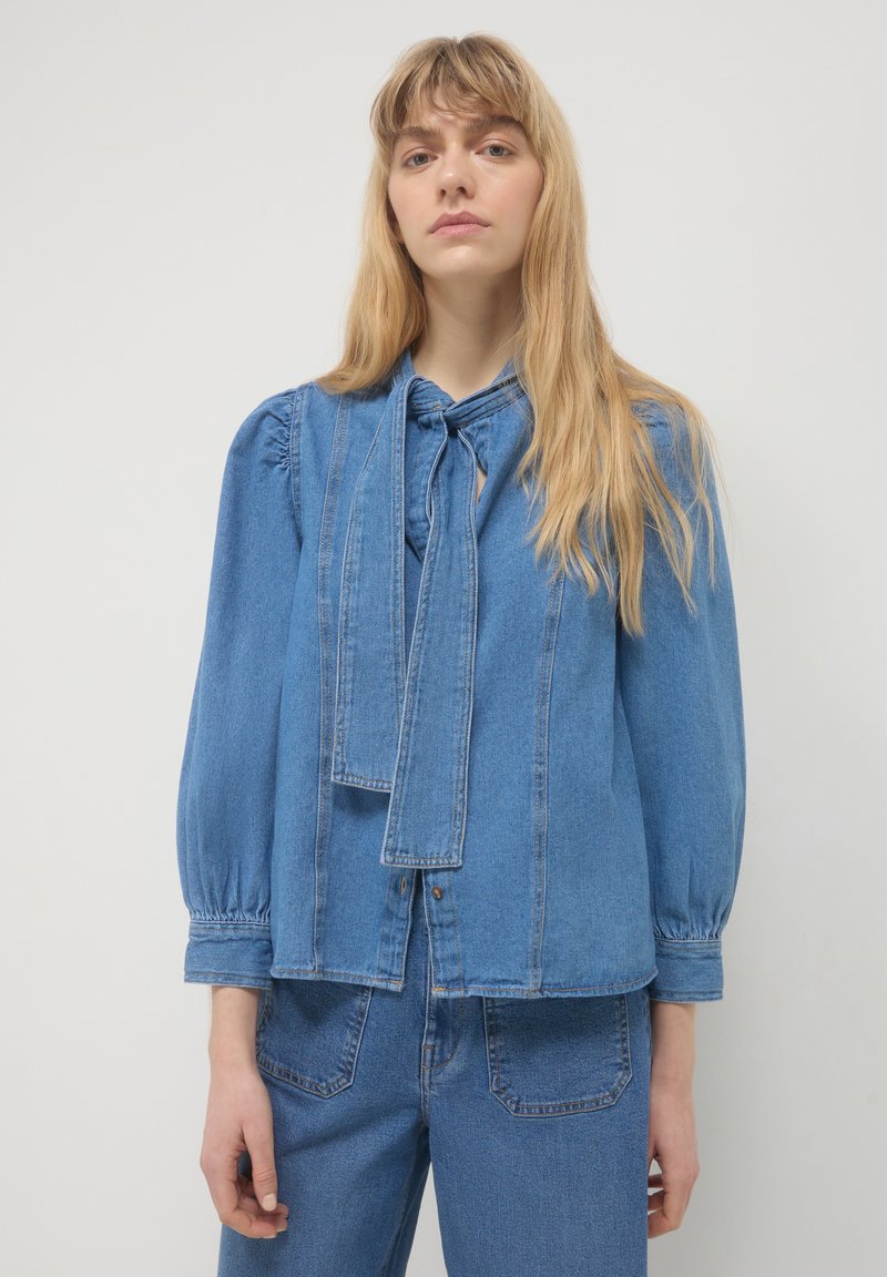 Femme aux longs cheveux blonds portant un chemisier en jean bleu avec un détail de col noué et des manches bouffantes, associé à un jean bleu.