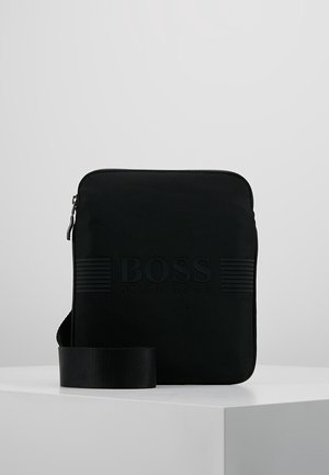 Musta neliönmuotoinen olkalaukku vetoketjulla, kohokuvioitu "BOSS HUGO BOSS" -logo ja säädettävä hihna, esillä valkoisella pinnalla.