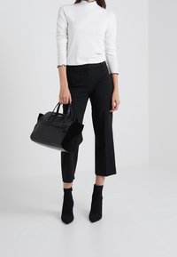 Sac à main noir avec une surface texturée, doté de poignées lisses et d'accents en daim contrastants. Le mod èle porte un haut blanc côtelé et un pantalon noir.