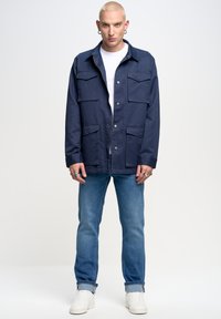 Giacca leggera blu navy con maniche lunghe, numerose tasche frontali e taglio dritto, abbinata a jeans azzurri e sneakers bianche.