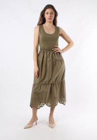 Abito senza maniche di colore verde oliva con un design a strati; il bordo in pizzo presenta ritagli floreali. Indossato con tacchi nudi.