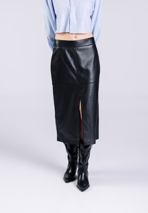 Falda midi de cuero negro con una abertura frontal, textura suave y cintura ajustada. Combinada con botas negras de punta afilada que presentan adornos decorativos.