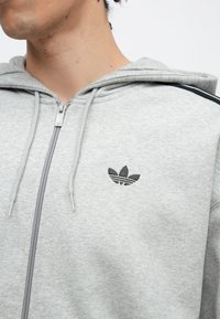 Jovem vestindo uma camisola com fecho éclair cinza claro com o logótipo preto da trevo da Adidas no peito e uma risca preta no ombro.