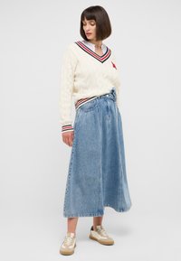 Ivory Pullover mit Kabelmuster und V-Ausschnitt, ausgestattet mit roten und navyfarbenen Streifen. Fließender, hellblauer Denim-Maxirock mit gebundenem Taillenbund. Beigefarbene Sneakers.