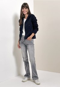 Felpa con zip blu navy e maniche testurizzate, t-shirt bianca, jeans a campana grigi chiari e sneakers bianche; in piedi contro uno sfondo chiaro.